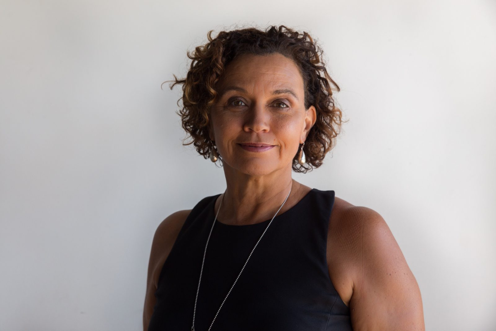 Portrait of a middle-aged woman with short curly hair wearing a black sleeveless top and silver jewelry, against a light background.